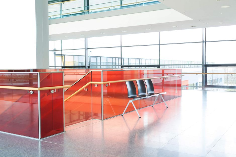 Powder coated red glass partitions in a staircase railing in a well lighted building with three black plastic seats right beside the end of the staircase