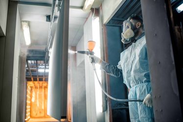 Man wearing a protective suit and respiratory mask is powder coating a metal sheet in a spray booth holding a powder gun in his right hand