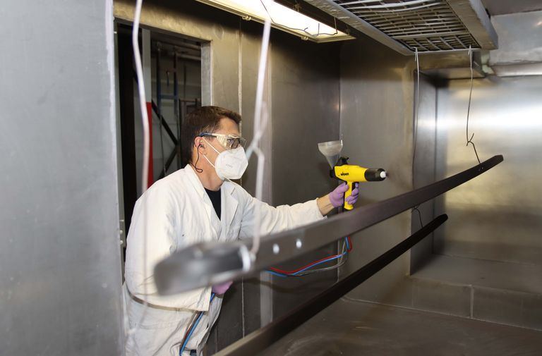 Man powder coating roof rack bars black inside of a powder coating booth while wearing a lab coat, breathing mask and protective glasses